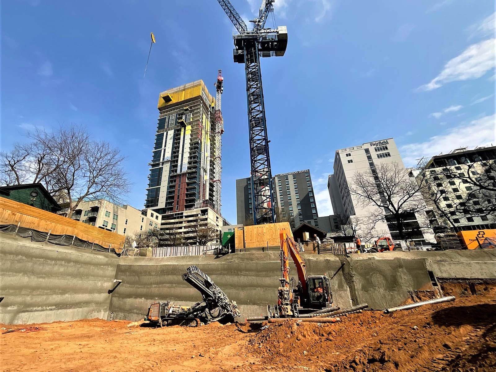 Compact drilling rig and excavator from Oscar Orduno, Inc. installing Anchored Soldier Piles and Soil Nail Anchors at Paseo 80 Rainey in Austin, TX.